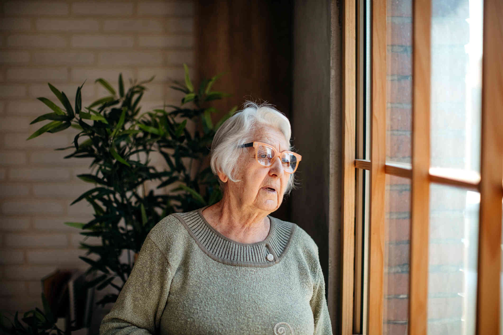 An elderly woman in a green sweater stands near a window in her home and gazes out while looking confused.